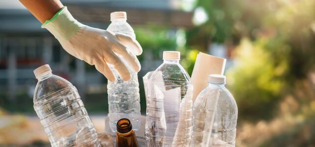 A gloved hand places a plastic bottle into a cardboard box filled with empty plastic and glass containers, highlighting efforts for plastic recycling in Singapore outdoors.