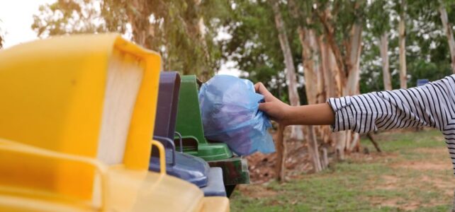 A person in a striped shirt places a blue plastic bag into a green recycling bin outdoors near trees, practicing eco-friendly junk pick up.