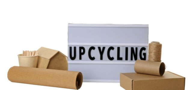 A sign reading "UPCYCLING" sits among cardboard, paper, a small upcycled cardboard house, twine, and recyclable containers, all on a white background.