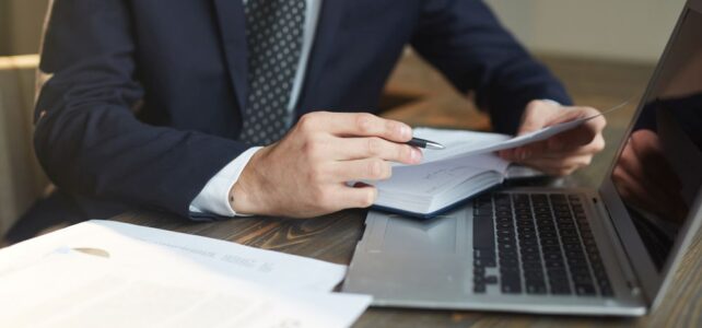 A person in a suit reviews documents while using a laptop at a desk with paperwork, ensuring compliance with 468 rule guidelines.