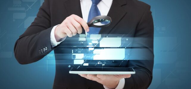 A man in a suit holds a magnifying glass over a tablet device, with digital interface graphics appearing above the screen, symbolizing the precision and focus of technology executive search.