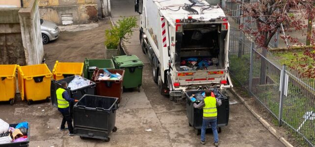 Two sanitation workers in yellow vests empty large bins into a garbage truck in an urban alley with several dumpsters nearby, highlighting the efficiency of junk disposal services.