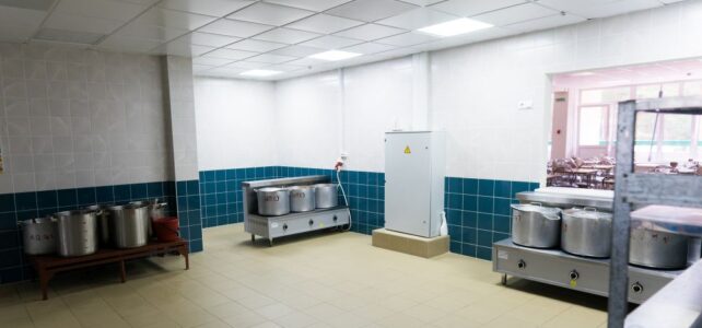 A commercial kitchen in an industrial canteen for rent, featuring large metal pots on stoves, a white storage unit, tiled walls, and an open window to another room.
